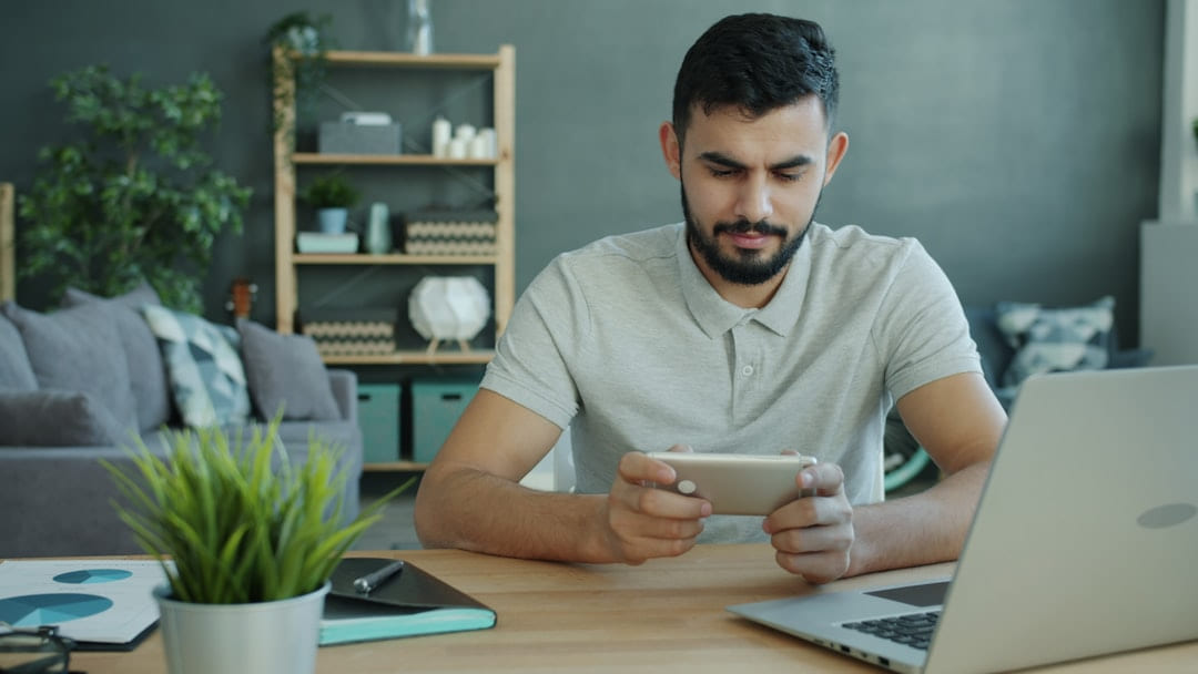 Man playing game on smartphone at desk with laptop on Affordable Smart Living